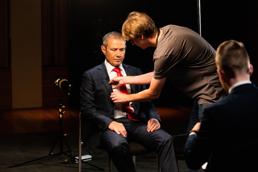 A man in a brown shirt adjusts a microphone on Roger Cook's suit jacket before an interview.