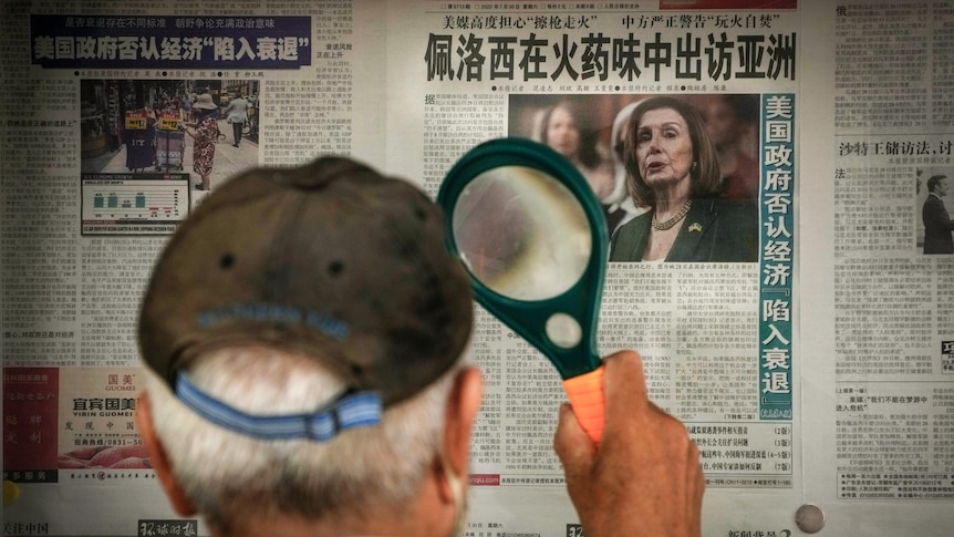 A man in a baseball cap, photographed from behind, holds a magnifying glass to a newspaper featuring Nancy Pelosi