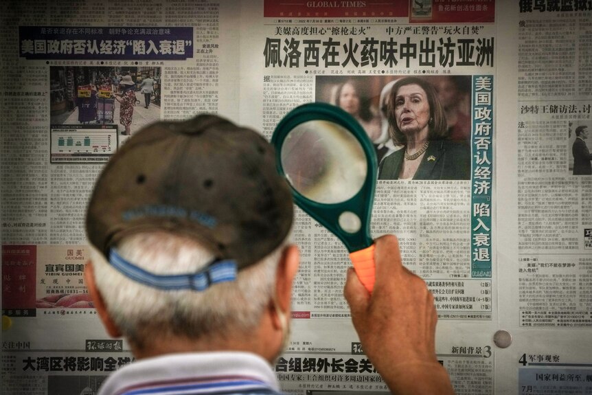 A man in a baseball cap, photographed from behind, holds a magnifying glass to a newspaper featuring Nancy Pelosi