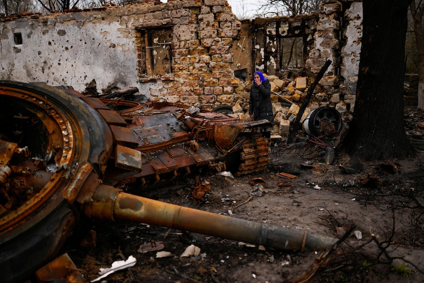 A woman stands next to a Russian tank in the backyard of her father's home, both destroyed.