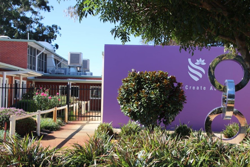 A school campus with buildings and shrubs and trees with a purple wall in the background and a sculpture.