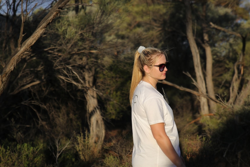 A young woman with a blonde hair and a white tshirt stands in bushland.