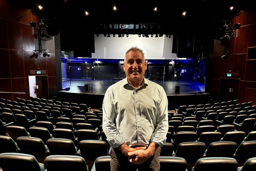 Man stands in front of rows of chairs with theatre stage behind him.