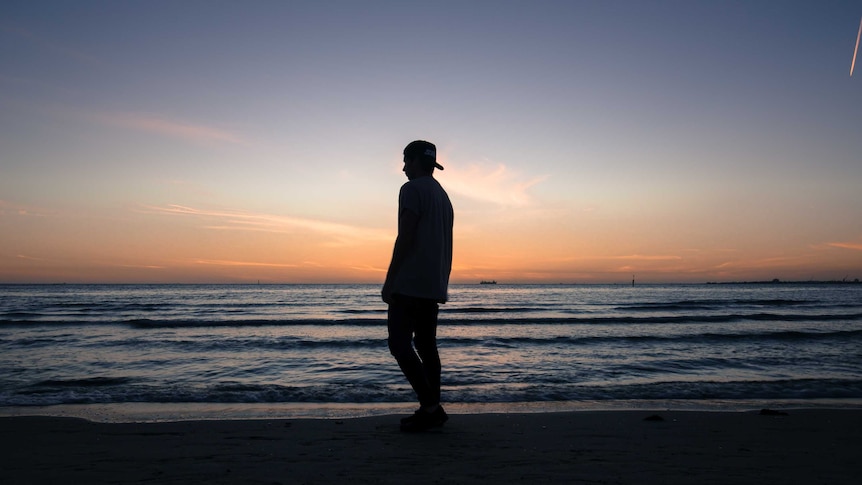 A man in a baseball cap on backwards stands by a beach.