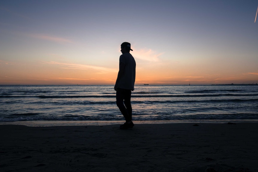 A man in a baseball cap on backwards stands by a beach.