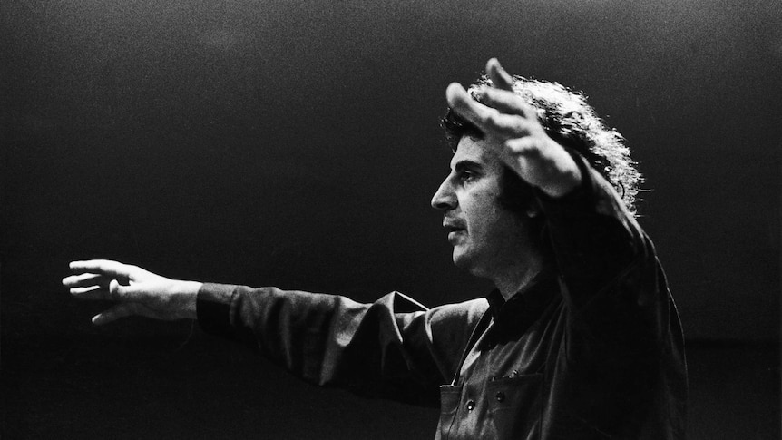 A black and white image shows Mikis Theodorakis conducting an orchestra in profile
