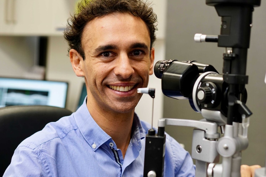 A smiling Kristopher Rallah-Baker sits in front of eye testing equipment.