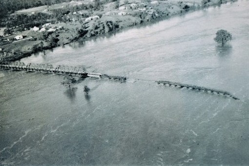 Black and white aerial image shows river lapping at bridge and some sections washed away.