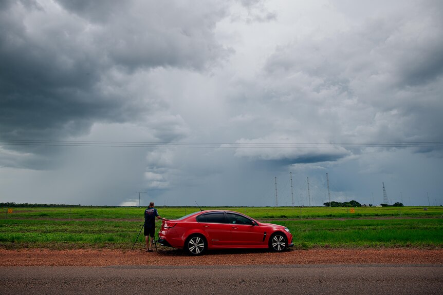 A man stands next to a red car on the side of a rural road. 