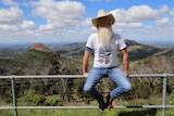 Man with big beard and hat sits on fence with mountain range behind