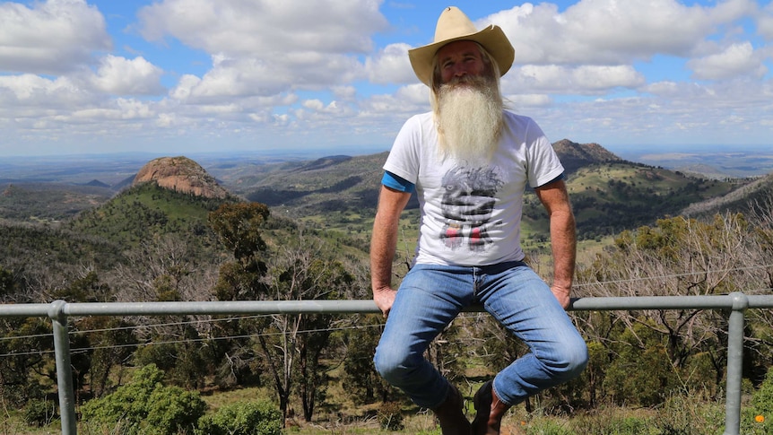 Man with big beard and hat sits on fence with mountain range behind