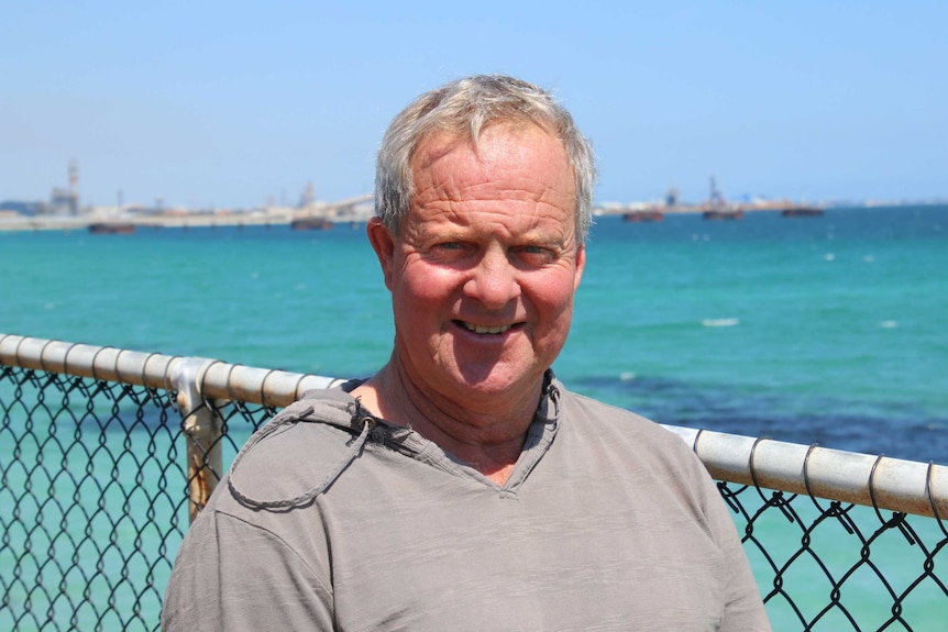 A smilig man wearing a loose grey top standing behind a fence with an aqua blue ocean in the background