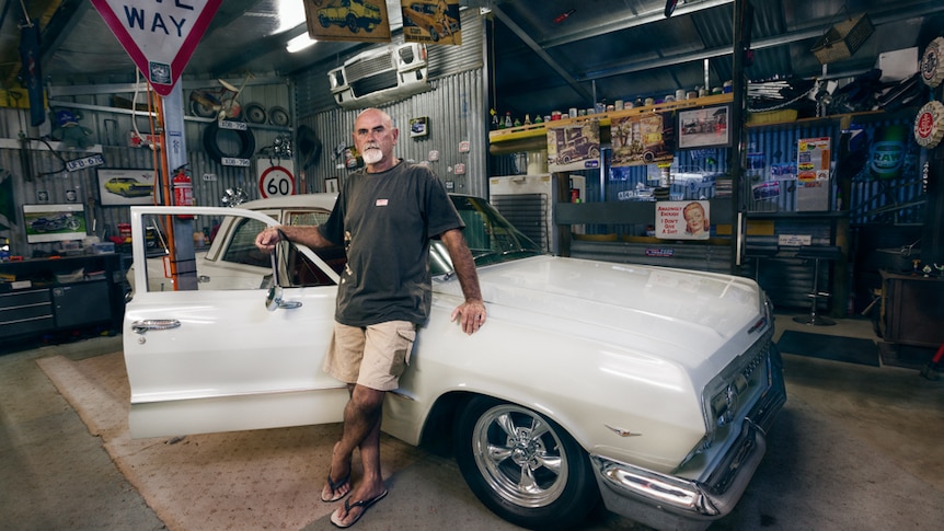 A man leans against a vintage car in a garage filled with automobile tools and paraphernalia.