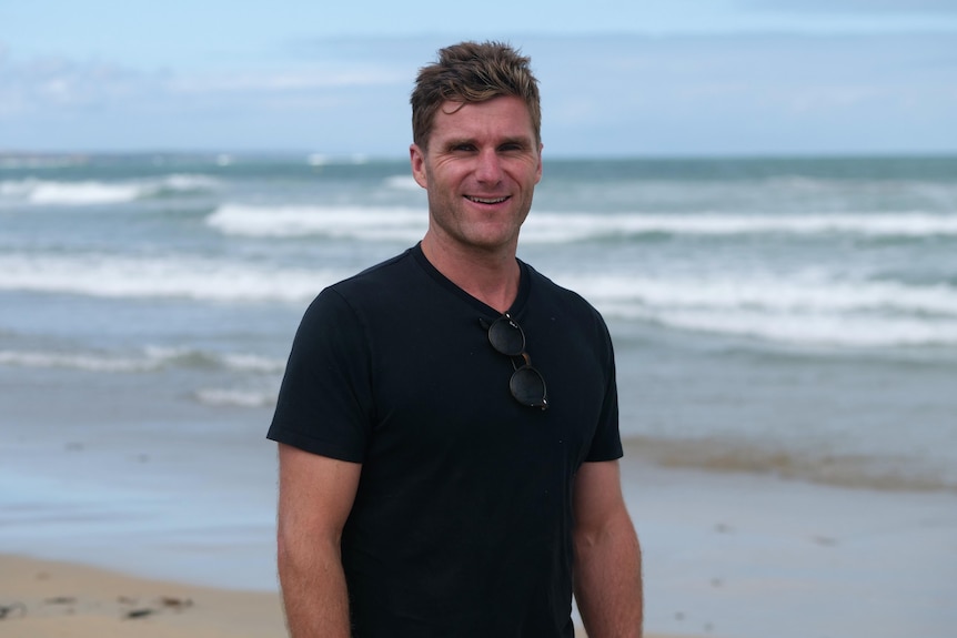 Fit young man stands smiling on the beach wearing black t-shirt