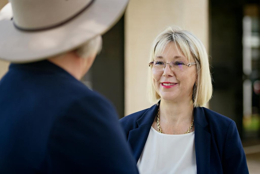 A medium close-up of a blonde woman in glasses a blue blazer speaking to someone in a hat.