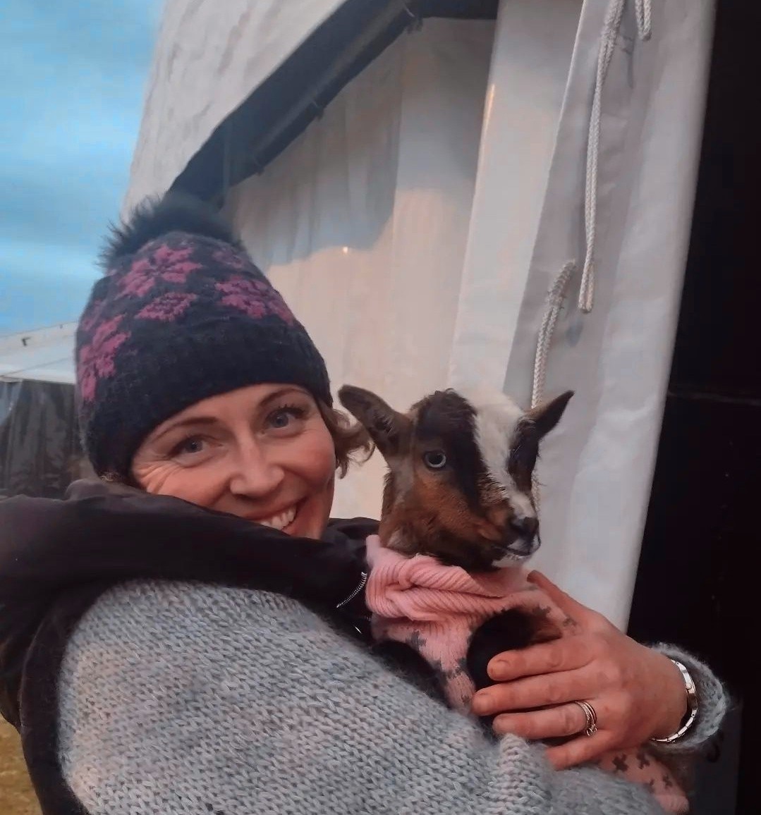 A woman wearing a purple and black woollen beanie, holding a small brown and white goat wearing a pink scarf.