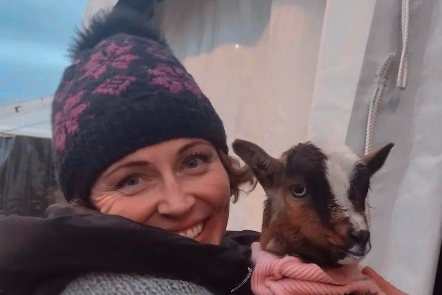 A woman wearing a purple and black woollen beanie, holding a small brown and white goat wearing a pink scarf.