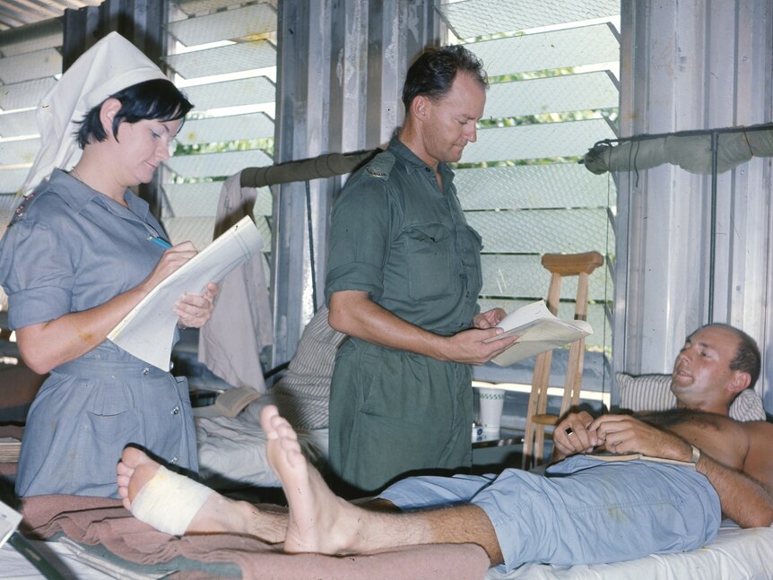 A female nurse and male medic stand by the bed of a male patient during the Vietnam War