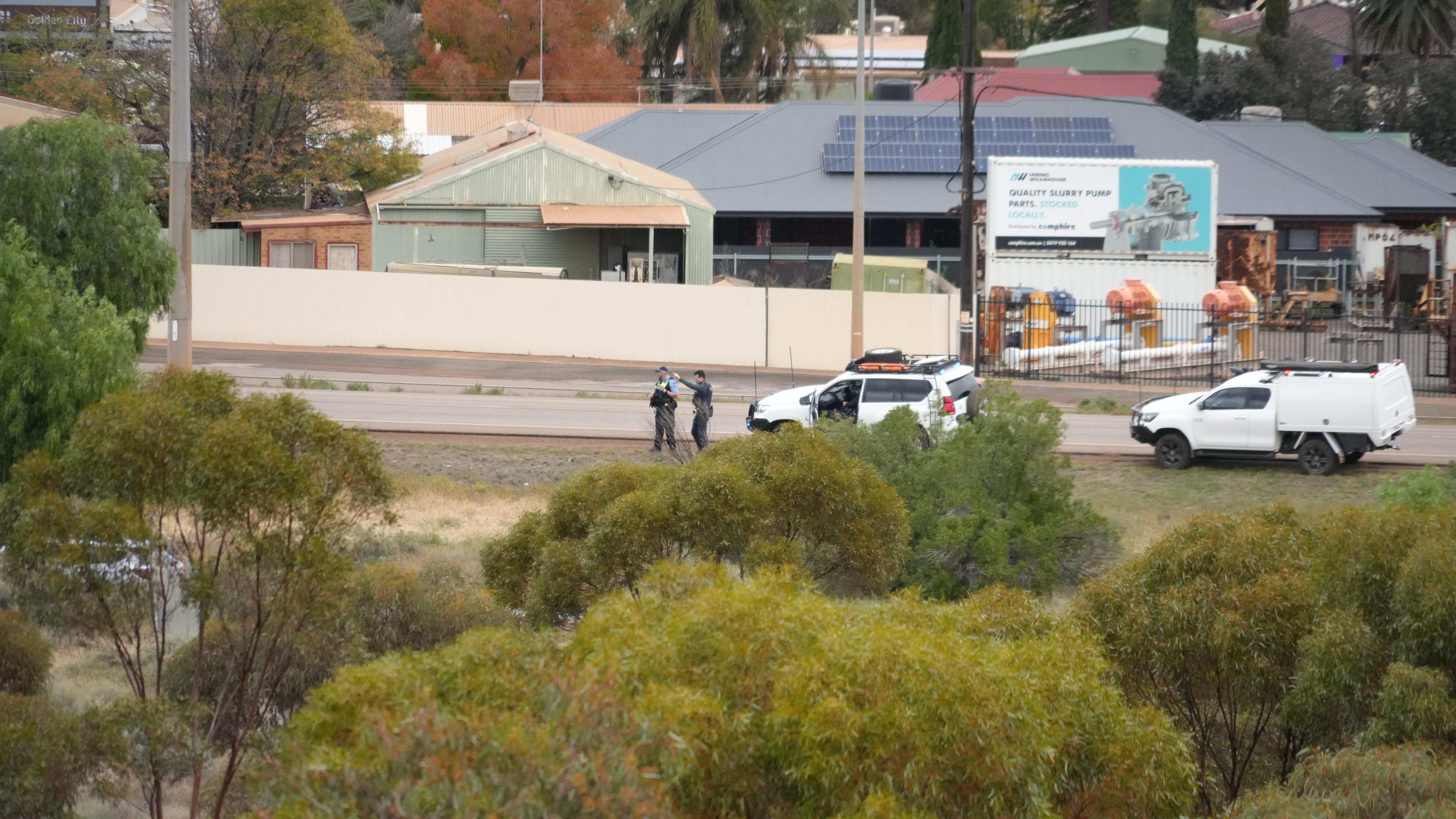 Two white cars parked at the side of a road with two men, one in a police uniform standing by them, bushland in the foreground