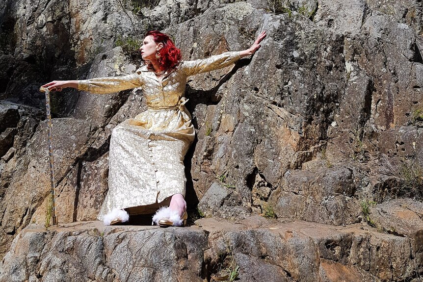 A colour image of Michelle, a white woman with red hair wearing a long, golden dress, on a rock face with a walking stick.