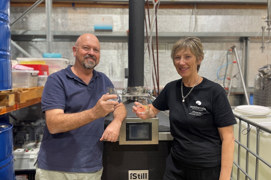 A middle-aged man and woman holding glasses of gin and smiling in front of steel still.