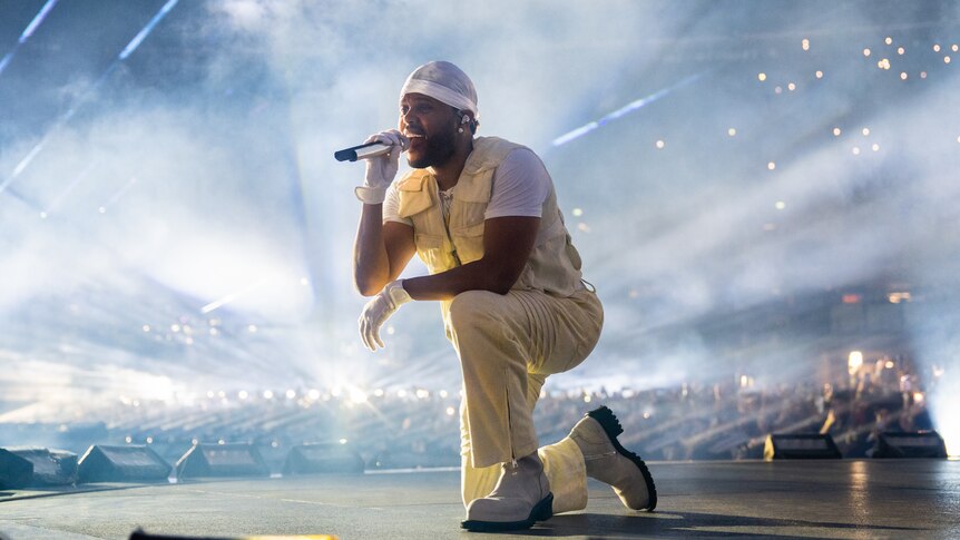 Abel Tesfaye aka The Weeknd kneels on a stadium stage in an all white outfit white singing into a microphone