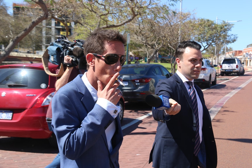 Mark Sebo walks while wearing sunglasses and smoking a cigarette as a reporter asks him a question
