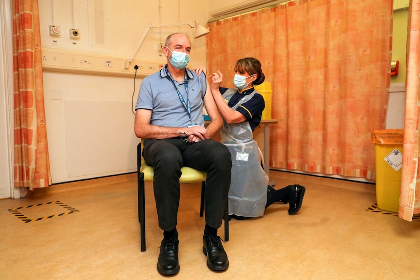 A man wearing a mask receives an injection in his arm from a woman wearing a mask.