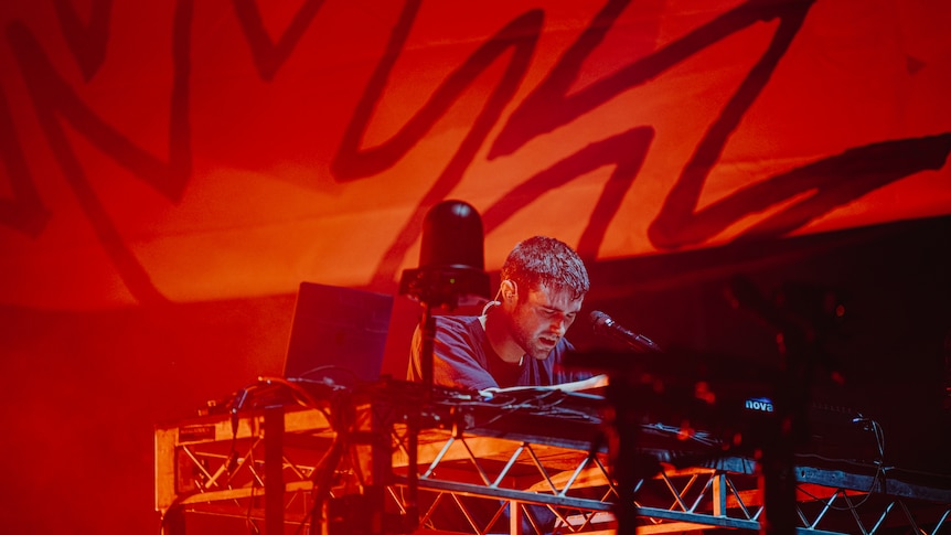 Fred Again performing at Laneway Festival hunched over DJ decks with red lighting behind him