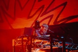Fred Again performing at Laneway Festival hunched over DJ decks with red lighting behind him