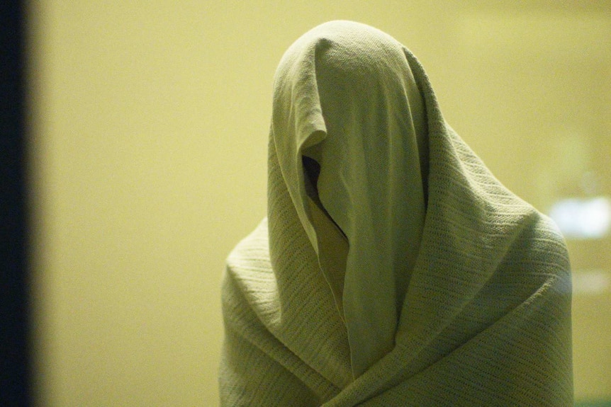 Prisoner inside a watch house stands with sheet draped over their head.