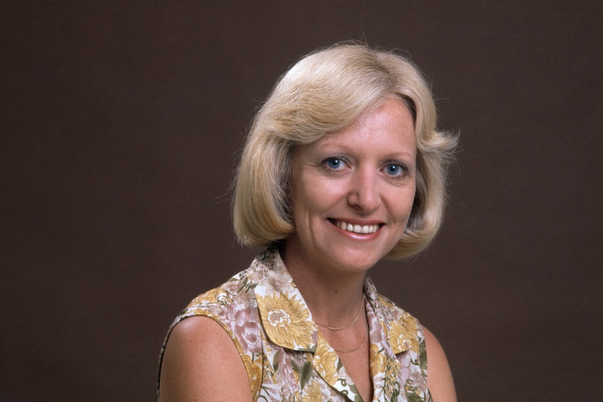 A young Kate Sullivan wearing a floral top, with blonde hair, smiling on a brown background.
