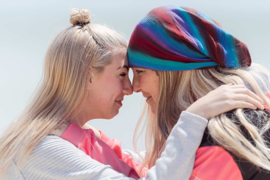 A close-up shot of a teenage girl and her mother smiling with their heads pressed against one another.