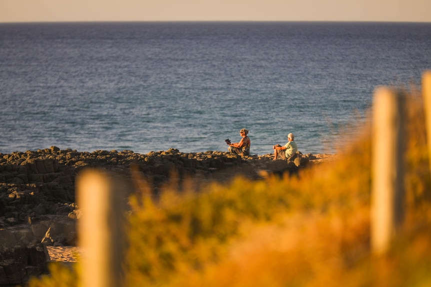 Two older women sit in fold-out chairs on rocks at the beach.