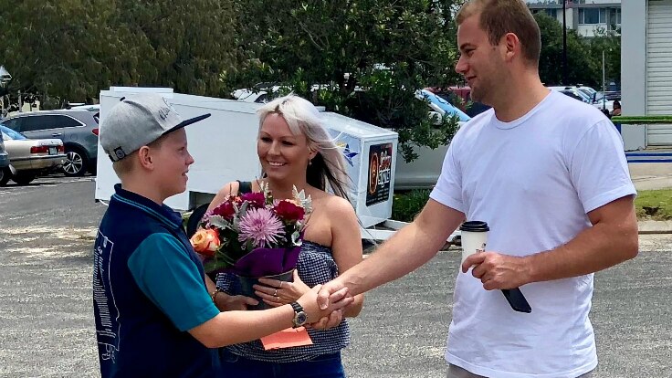 An 11-year-old boy shakes hands with a man who saved his life in the surf while mum looks on holding flowers.