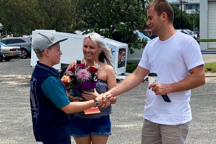 An 11-year-old boy shakes hands with a man who saved his life in the surf while mum looks on holding flowers.