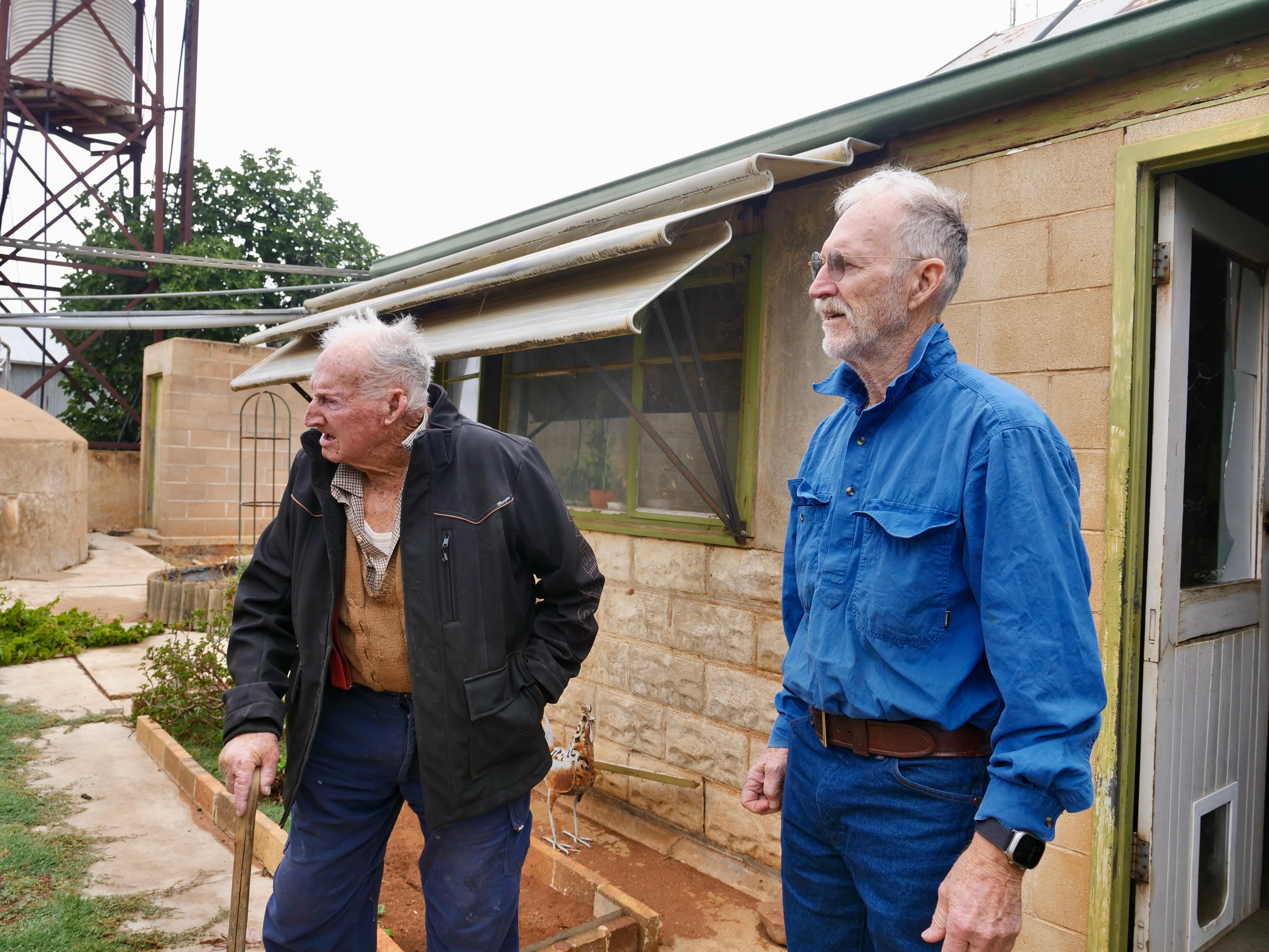 two men standing, one with a walking stick and a black jacket, one with a blue shirt grey hair and glasses looking away 