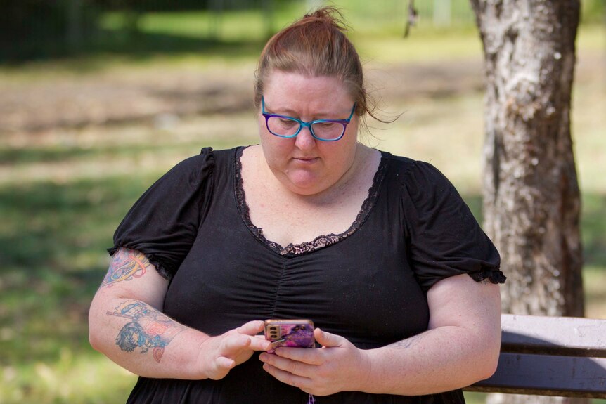 Sharmy is sitting down outside. She holds her mobile phone in both hands. She looks down at it.