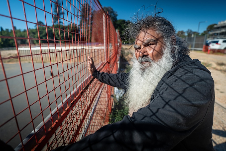 An elderly man with white beard looks at foundation on the ground surrounded by red fence