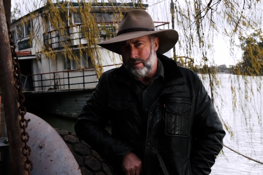 Man in hat by the Murray River in front of a paddleboat