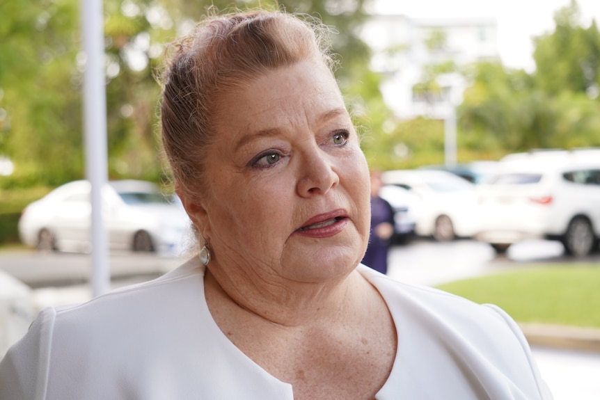 Sue Ellery wearing a white jacket, standing outside parliament on a rainy day, looking away from the camera.