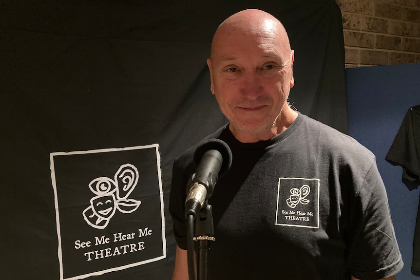 Craig Palmer wears a black shirt and stands next to the See Me Hear Me Theatre logo.
