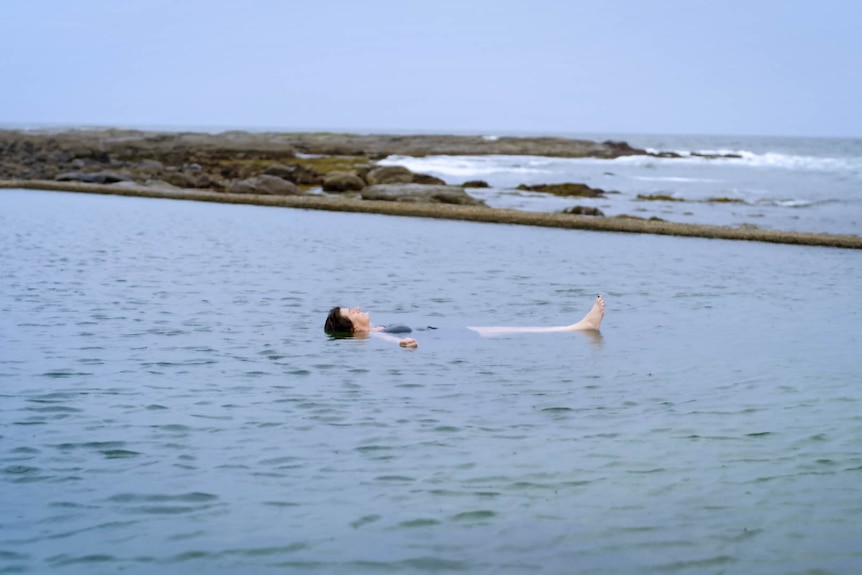 A woman floats on her back in an ocean pool on an overcast day.