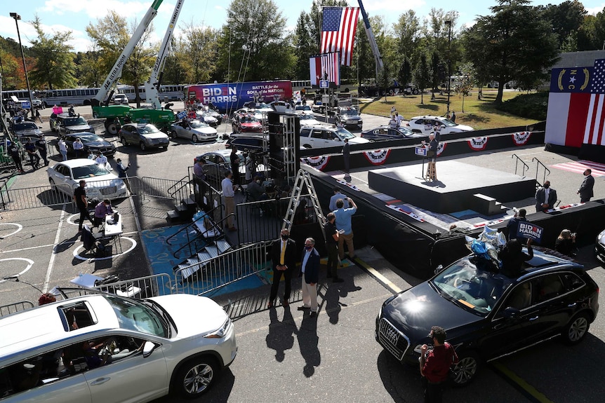 Joe Biden stands alone on a large stage set up in a car park surrounded by cars. Camera equipment and technical staff are nearby