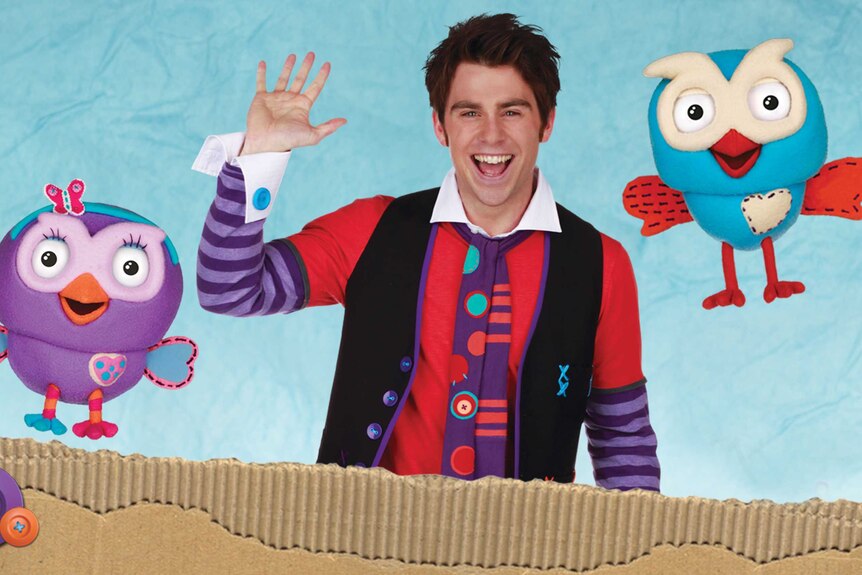 A man in brightly coloured clothing waves, flanked by two puppet owls