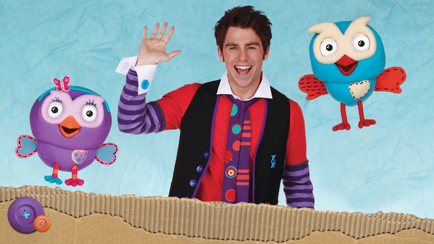 A man in brightly coloured clothing waves, flanked by two puppet owls