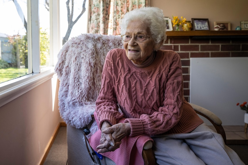 An elderly woman sits in a chair and looks out a window.