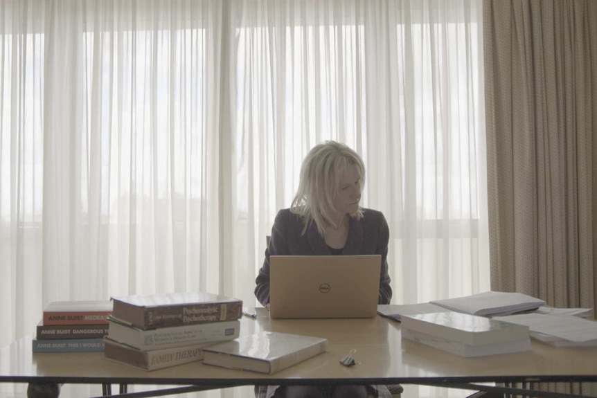 Woman on right of image looking at computer at desk in front of curtains