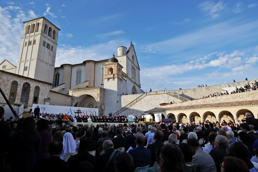 A large crowd sits in an open square and people stand along a wall to look down on a stage on which the Pope sits.