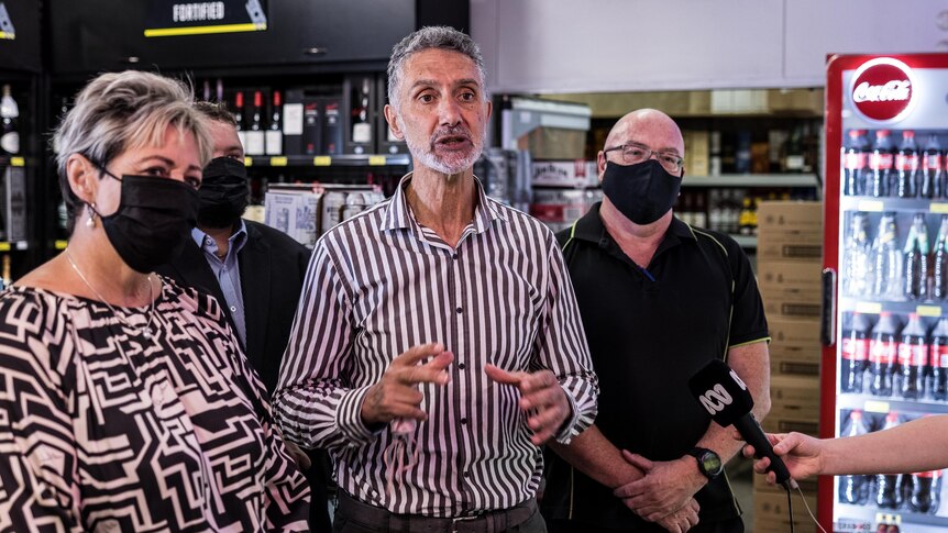 A male MP speaking to media inside a liquor store surrounded by people wearing masks.  